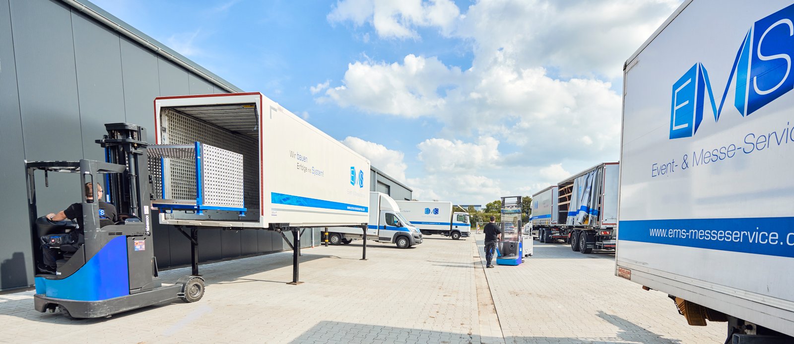 Gabelstapler belädt einen EMS-Anhänger mit modularen Messestand-Elementen vor einer Lagerhalle